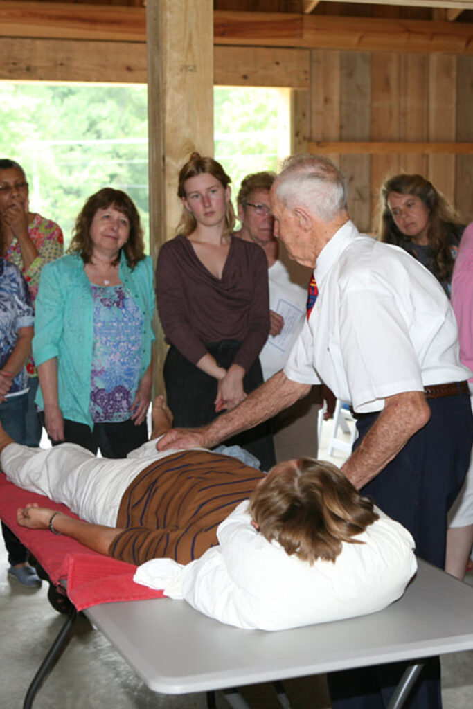 Geoffrey Morell demonstrating Clendenning Healing Technique on woman lying face up on massage table surrounded by students looking on